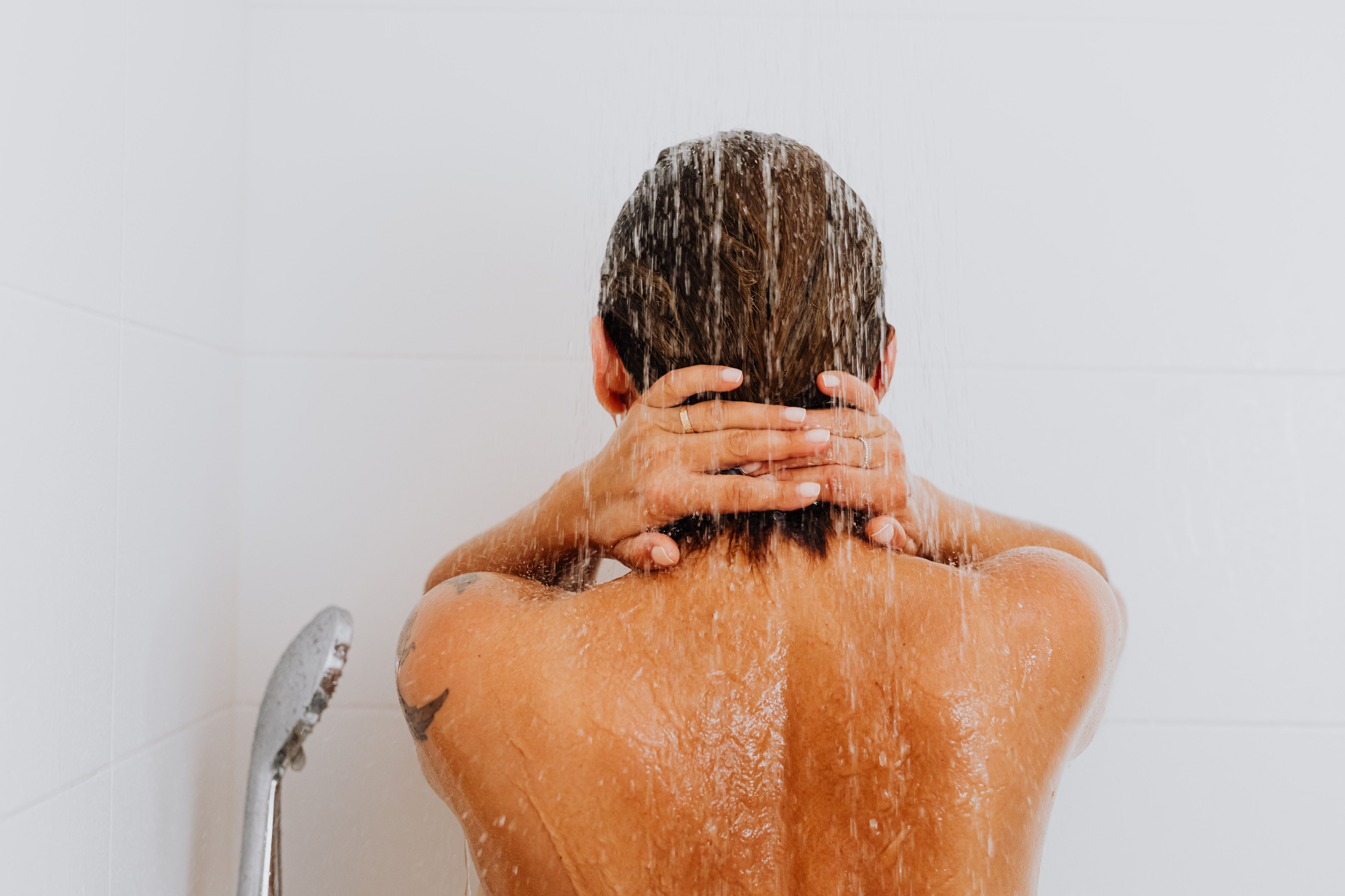 Person seen from behind under a running shower, water running over hair and back